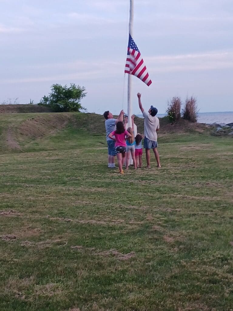 Family raising the American flag on a grassy field at Point Lookout State Park, Maryland, honoring tradition and creating lasting memories.