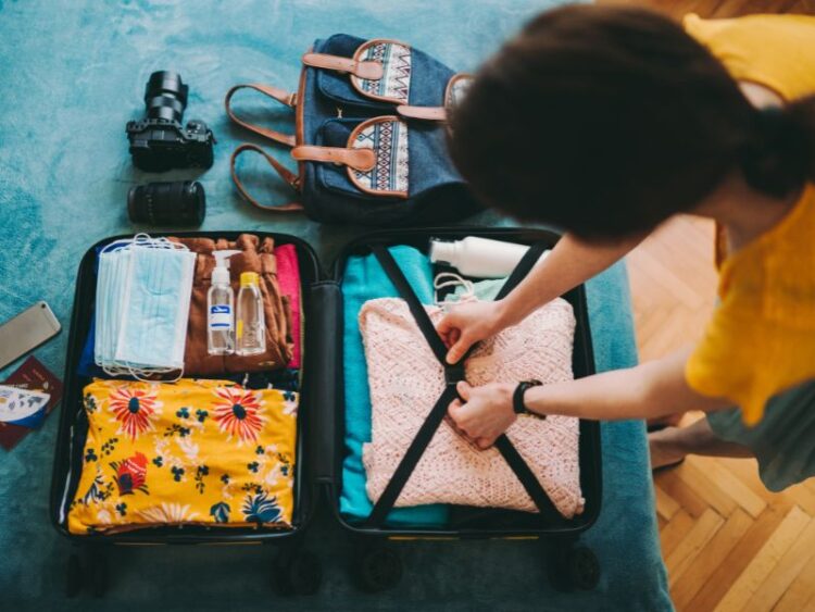 Traveler packing a suitcase with colorful clothing, face masks, hand sanitizer, and travel essentials, preparing for a trip with a camera, backpack, and passport nearby.