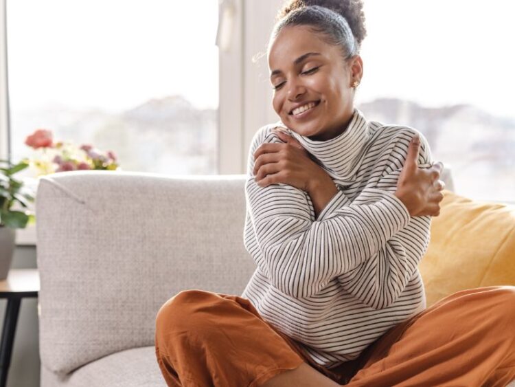 Smiling woman hugging herself on a sofa, practicing self-love and mindfulness at home.