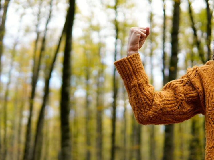 Person in cozy sweater raising fist in forest as a gesture of strength and empowerment