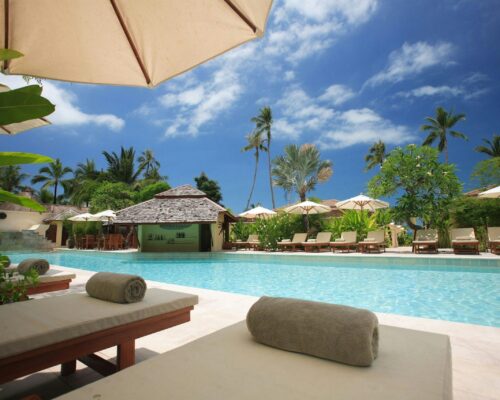 Experience ultimate relaxation at a sunlit tropical poolside resort featuring palm trees and blue skies.