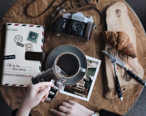 Flat lay of vintage camera, coffee cup, croissant, and journal on a wooden table, perfect for creative brunch vibes.