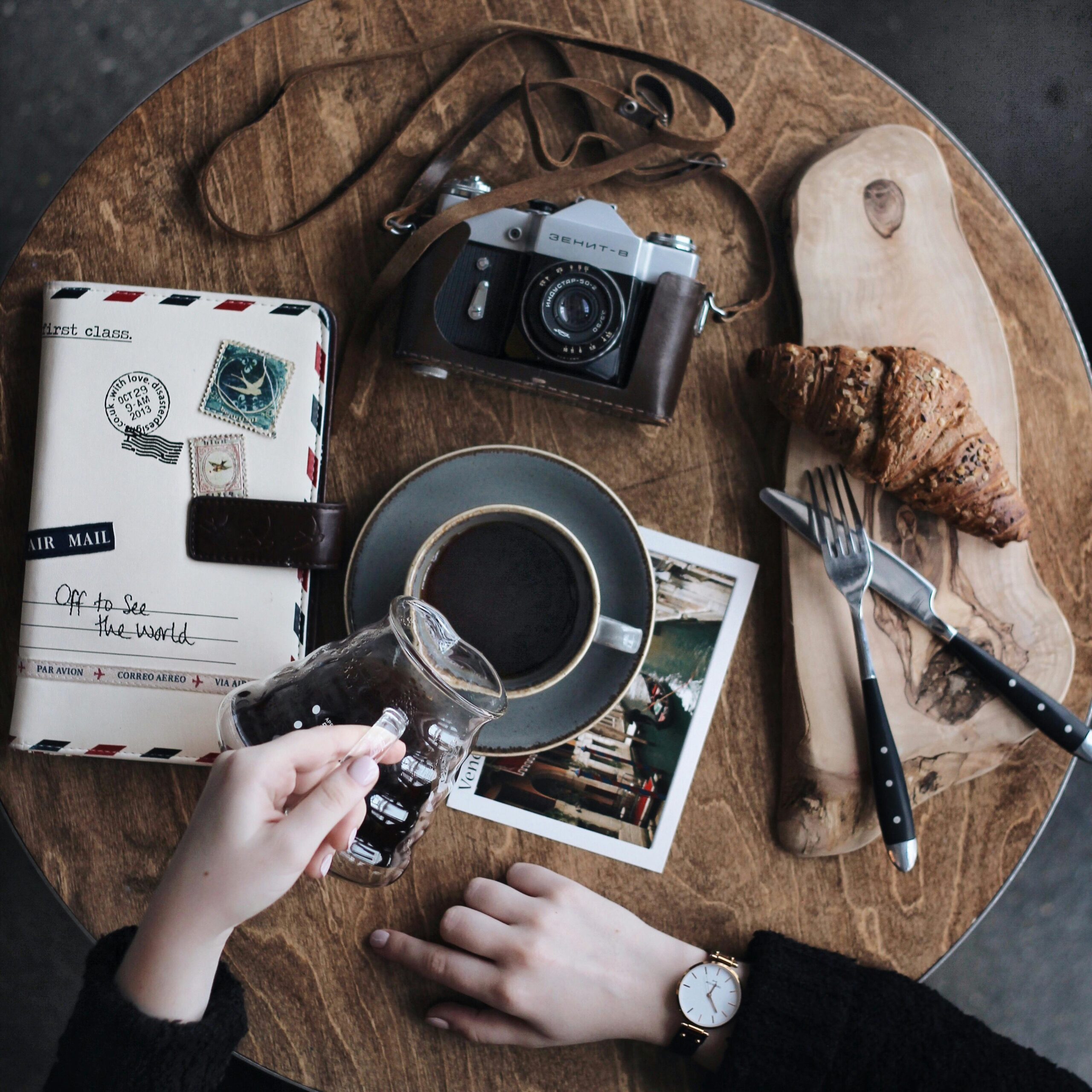 Flat lay of vintage camera, coffee cup, croissant, and journal on a wooden table, perfect for creative brunch vibes.