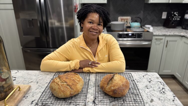 Woman smiling in kitchen with two fresh loaves of sourdough bread on cooling racks, illustrating mindful living with hands-on rituals.