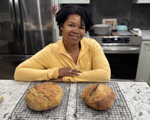 Woman smiling in kitchen with two fresh loaves of sourdough bread on cooling racks, illustrating mindful living with hands-on rituals.