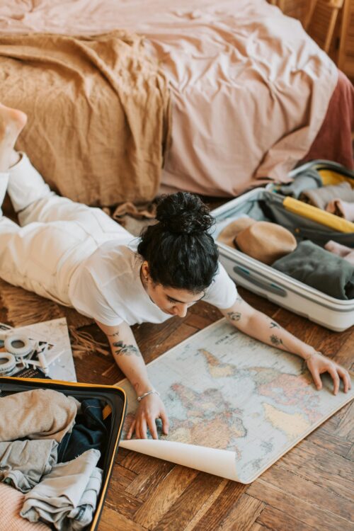 A woman lying on the floor indoors, with an open suitcase with light packing essentials laid out for a budget-friendly trip..