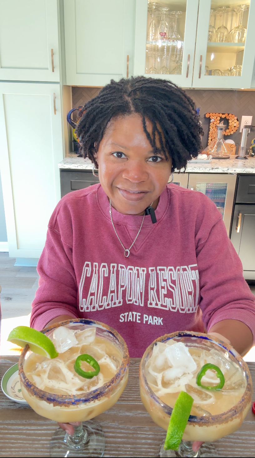 A woman in a Cacapon Resort sweatshirt holds two glasses of mindful Dark Chocolate Mexican Margarita with ice, a cocoa-chili rim, lime wedges, and jalapeño slices, set in a bright home kitchen.