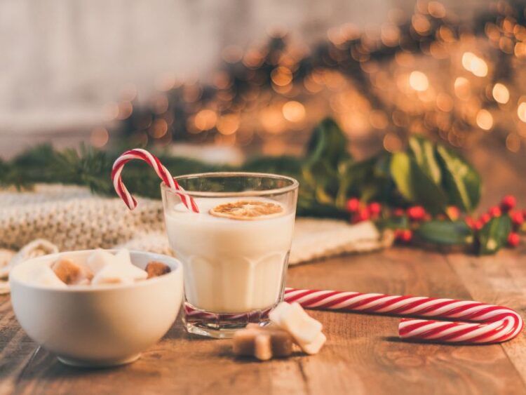 Holiday drink with a candy cane beside a bowl of marshmallows on a wooden table with festive decor in the background