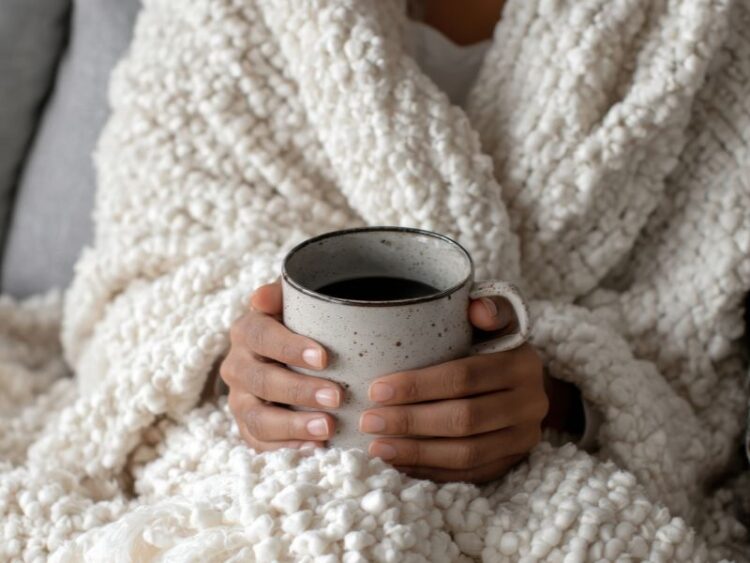 Hands holding a warm mug while wrapped in a soft blanket, representing mindful living and creating a calm, cozy moment during the holiday season.