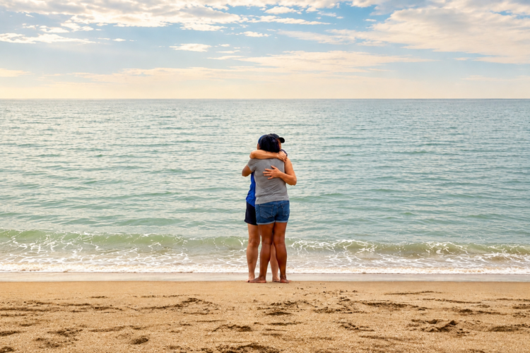 Two friends embracing on the beach while one gently rubs the other's back in a quiet moment of comfort and support.