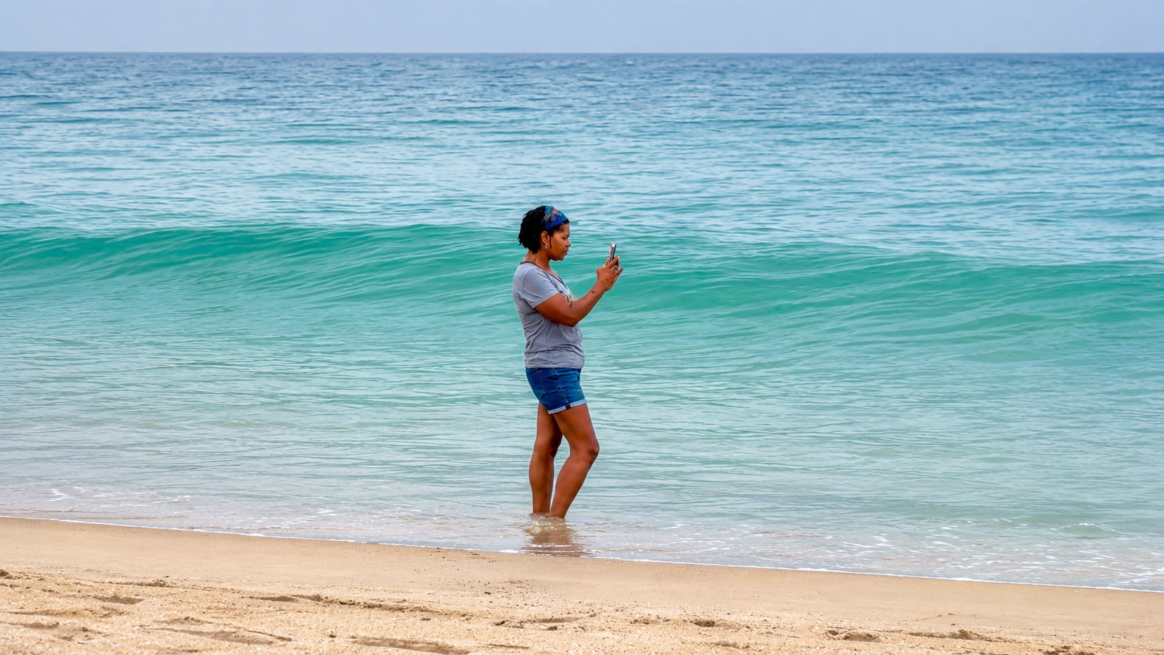 A woman standing in the ocean at the beach while quietly filming the waves during a reflective moment of calm.