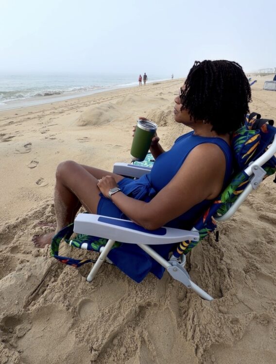 Woman sitting quietly in a beach chair with a cup of coffee, taking a peaceful morning pause by the ocean as a reminder that self-care doesn’t require hours to make a difference.