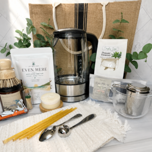 Even Here First Night Gift featuring Breville glass electric kettle, organic herbal tea, tea accessories, shower steamer, eye pillow, and new home essentials in a jute gift bag.