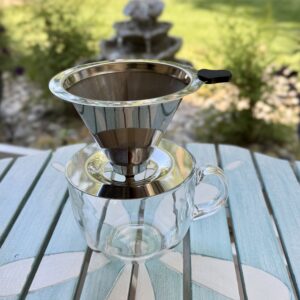 Reusable stainless steel pour-over coffee dripper resting on a clear glass mug outdoors with a softly blurred garden fountain in the background.