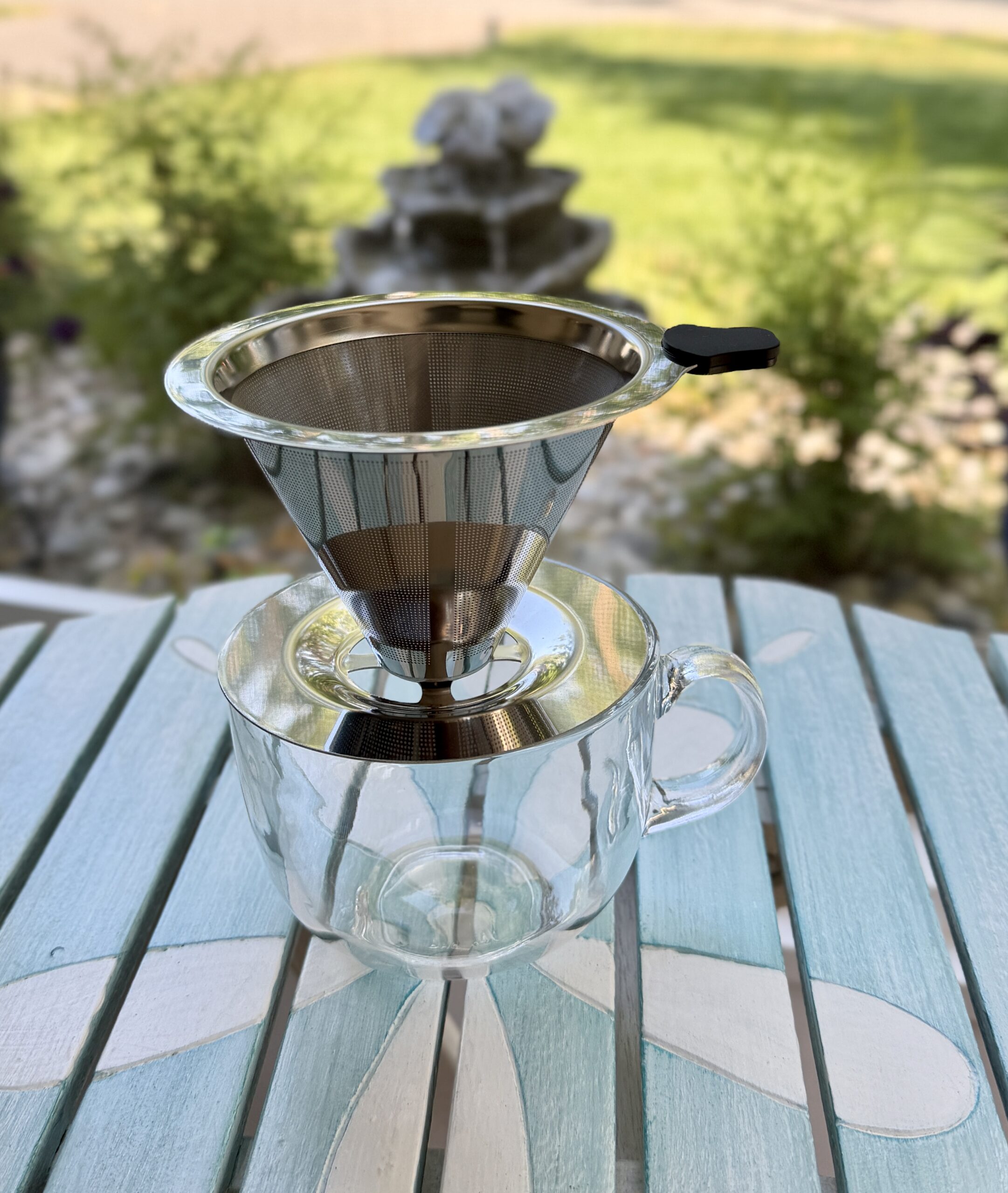 Reusable stainless steel pour-over coffee dripper resting on a clear glass mug outdoors with a softly blurred garden fountain in the background.